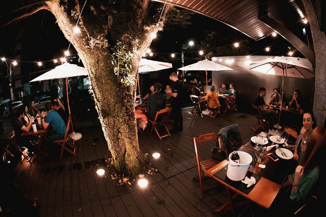 Foto noturna mostra o ambiente externo do Faro Cinco, em Criciúma. A imagem mostra um deck de madeira montado ao redor de uma árvore. Dezenas de pessoas estão sentadas em cadeiras de madeira ao redor de mesas de madeira, com ombrelones. Há um varal de luzes iluminando o ambiente. 