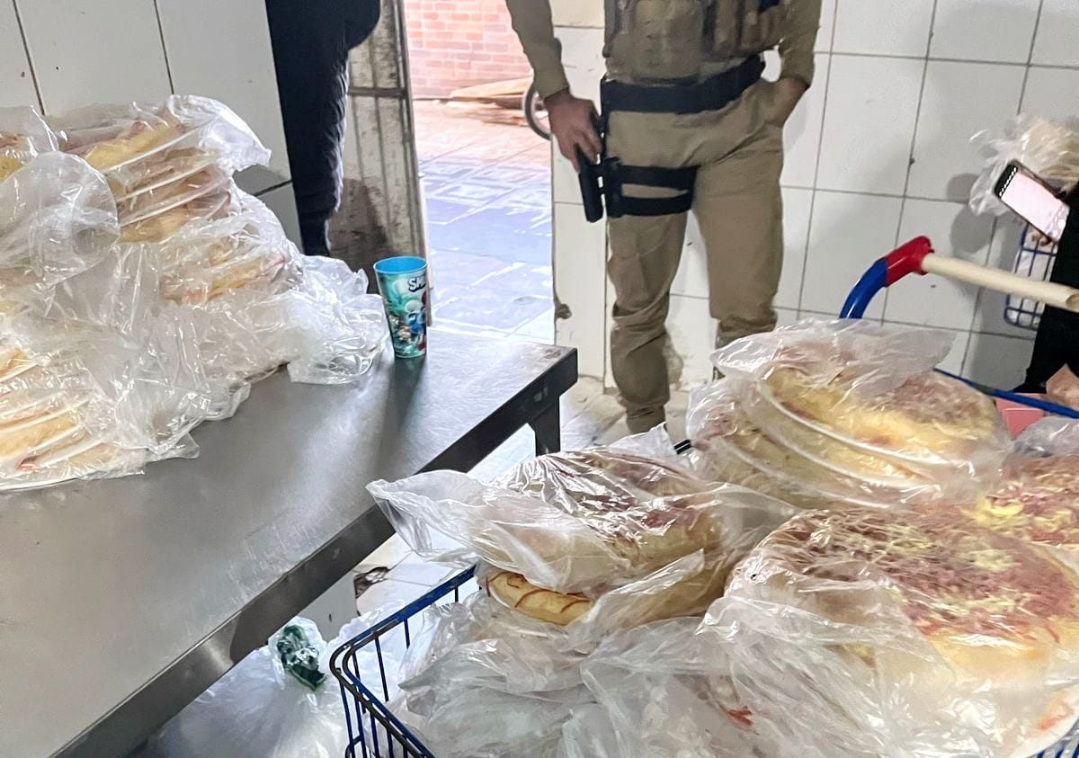 Foto mostra produtos apreendidos na fábrica de pizzas clandestina em Camboriú, com um policial militar ao fundo