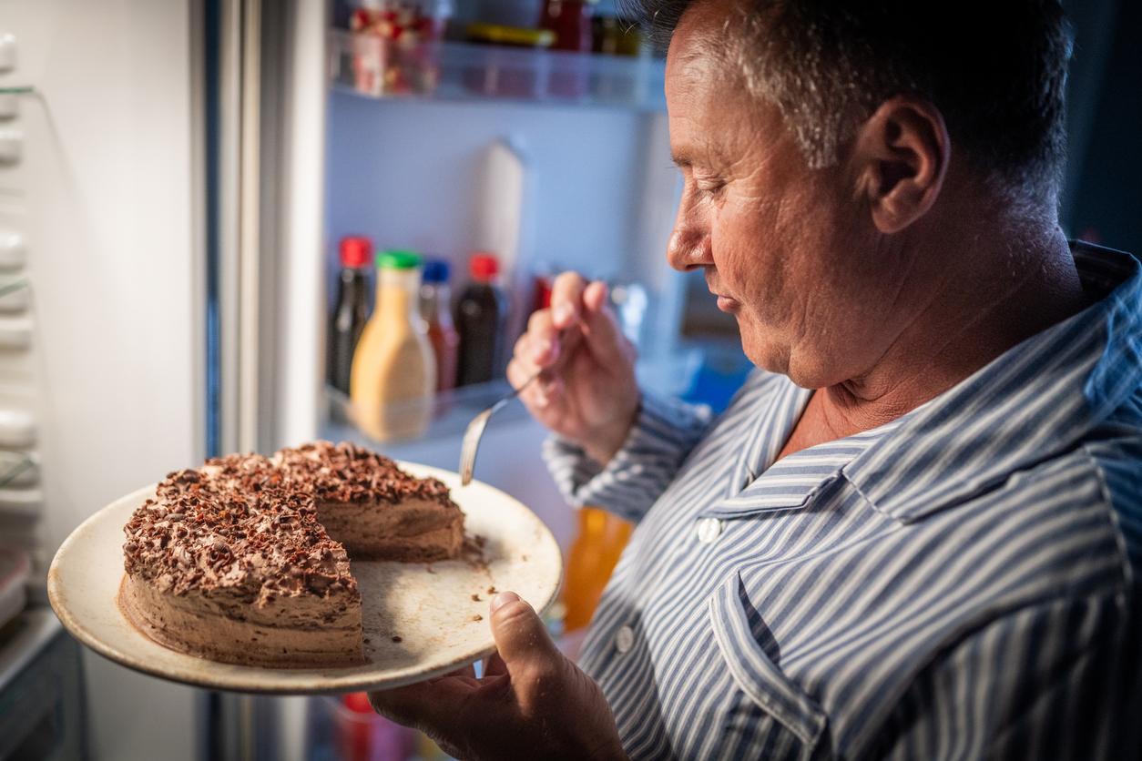 Homem comendo bolo em frente à geladeira no meio da noite para matar a vontade de comer doce
