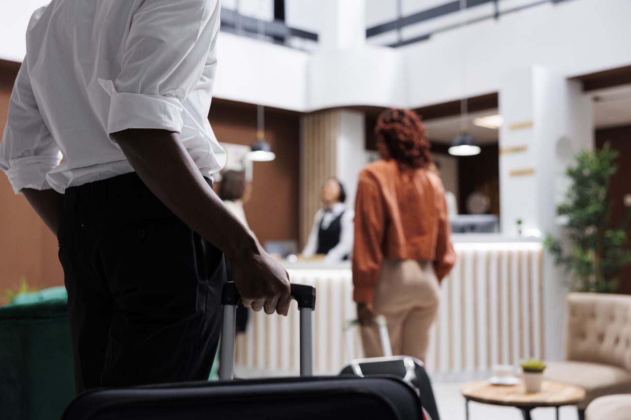 Homem carregando malas no saguão do hotel, chegando ao balcão da recepção com uma mulher para fazer o check-in. Hóspedes se preparando para preencher os formulários de registro, enquanto funcionários recebem os turistas