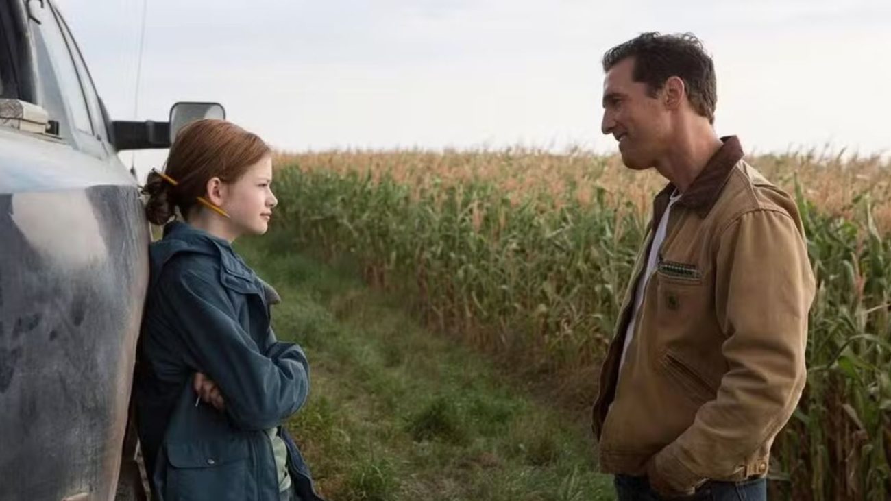 "Interestelar" está entre os filmes com finais surpreendentes que você precisa assistir. Na imagem, retirada do longa-metragem, dois personagens — um homem de cabelo escuro e jaqueta marrom e uma menina ruiva de jaqueta azul apoiada em uma caminhonete — conversam em frente a um milharal.