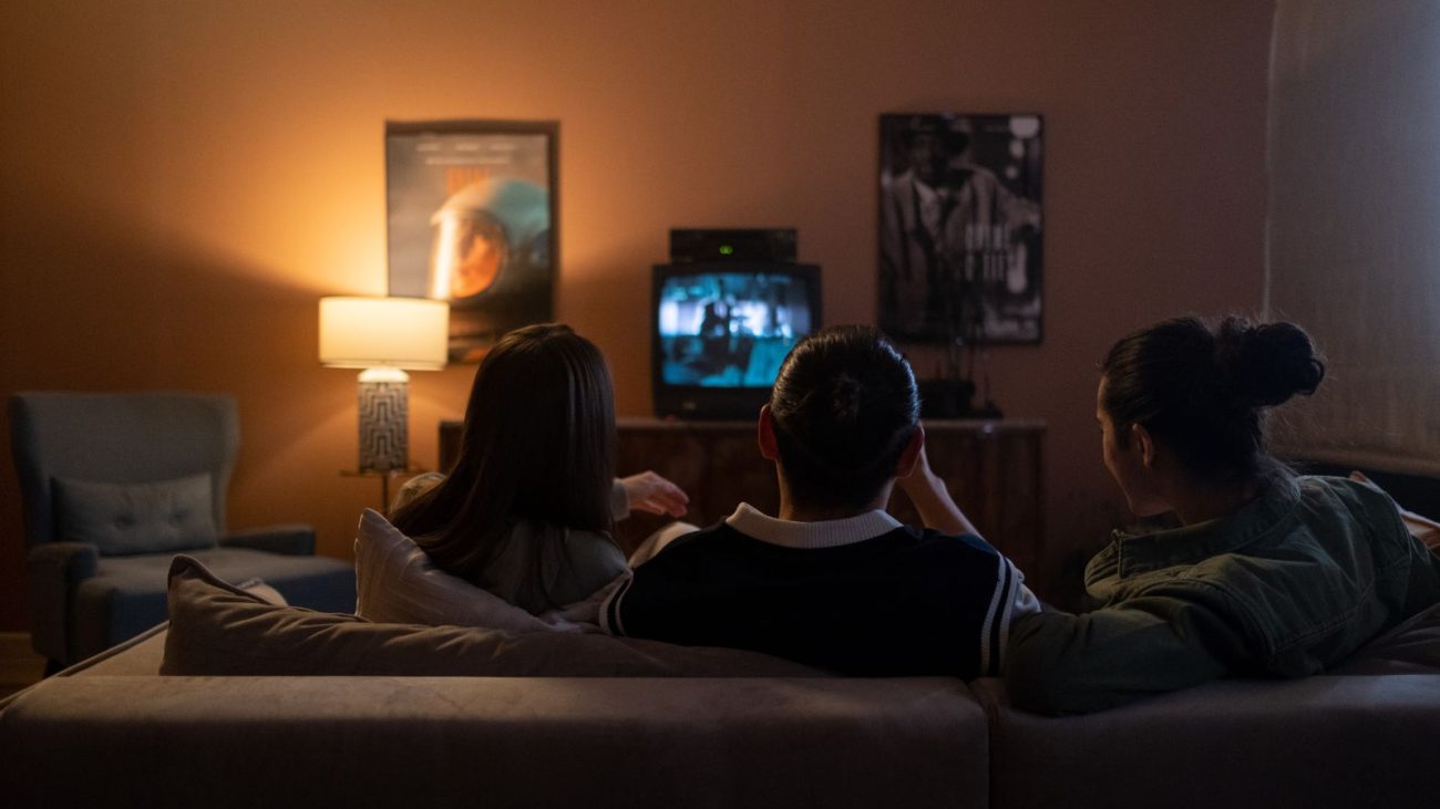 A imagem mostra três pessoas sentadas em um sofá assistindo a um filme em uma televisão. A cena transmite um clima aconchegante e intimista, com iluminação suave proveniente de um abajur à esquerda. As pessoas estão de costas para a câmera, de modo que seus rostos não são visíveis, e parecem estar envolvidas no que estão assistindo. A TV exibe uma cena em preto e branco, o que pode indicar que estão vendo um filme clássico. O ambiente tem uma decoração retrô: além da televisão antiga com videocassete, há pôsteres de filmes nas paredes e uma poltrona azul com almofada ao lado do abajur. É o tipo de imagem que remete a uma noite de cinema em casa, perfeita para curtir com a família ou amigos.