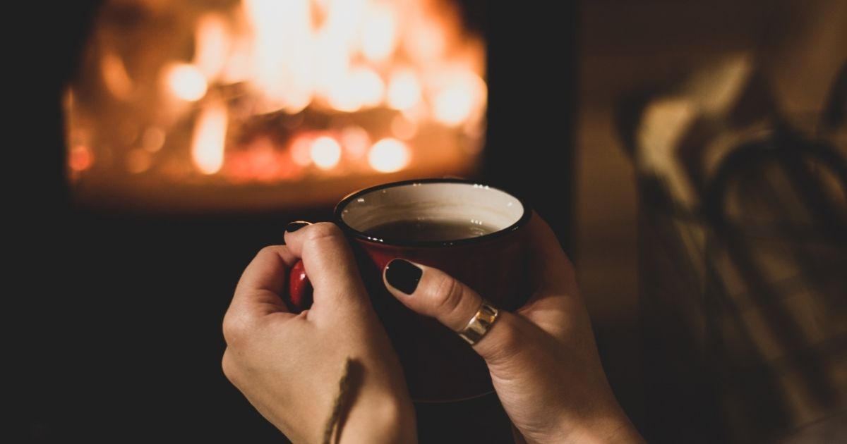Imagem de uma lareira com uma mão segurando uma caneca, para fazer referência ao fato de que as noites dessa semana terão temperaturas amenas 