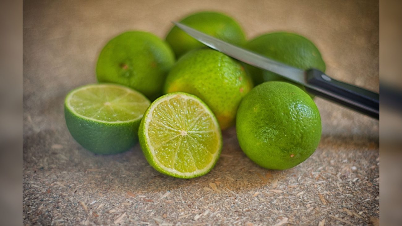 Vários limões verdes dispostos sobre uma superfície rústica. Dois deles estão cortados ao meio, revelando o interior suculento. Ao lado, uma faca com cabo preto está parcialmente apoiada sobre uma das frutas inteiras