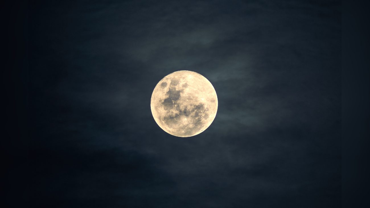 Fotografia de uma lua cheia brilhante no centro do céu noturno. A lua aparece com detalhes visíveis de sua superfície, como manchas escuras e crateras. Ao redor dela, o céu está escuro com nuvens finas e esfumadas, criando um clima misterioso e sereno