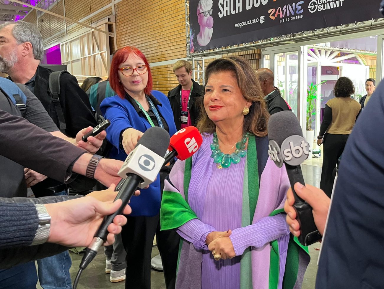 Luiza, de vestido roxo com verde, sorri enquanto conversa com jornalistas
