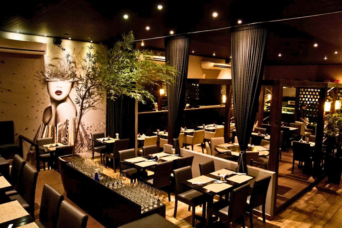 Ambiente interno com dezenas de mesas e cadeiras pretas. A luz é suave e ao fundo uma parede com a ilustração de parte de um rosto e corto de mulher. À frente da parede, uma planta alta com folhagens verdes. 