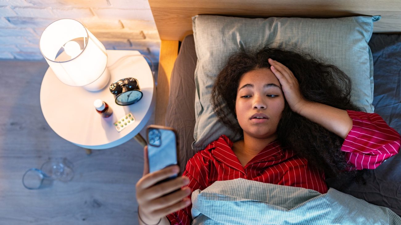 Mulher deitada na cama com expressão de cansaço, usando pijama vermelho listrado. Ela segura o celular com uma mão enquanto a outra está apoiada na testa. Ao lado da cama, há um criado-mudo com luminária acesa, um despertador, frasco de remédio, cartela de comprimidos e um copo com água no chão, sugerindo que ela está doente ou indisposta