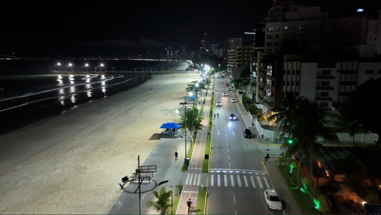 Foto noturna de Itapema. Do lado esquerdo, a areia e o meio. No meio, um calçadão com vários postes de luz. À direita na foto, uma via de asfalto por onde passam carros e onde estão alguns prédios.