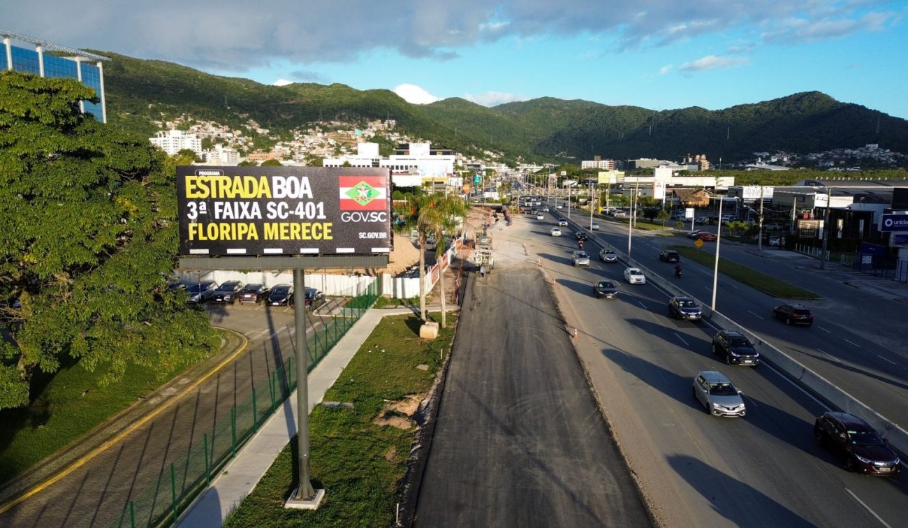 Vista aérea da SC-401 no Norte da Ilha, mostrando frentes de obra em andamento para a construção da terceira faixa pelo Programa Estrada Boa, com escavações e maquinário operando ao lado da pista existente cercada por mata nativa