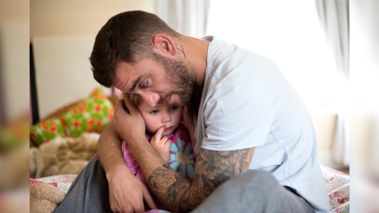 Homem com expressão preocupada abraça e conforta uma criança pequena, que também parece triste e está com o dedo na boca. Ambos estão sentados em uma cama, em um ambiente doméstico iluminado pela luz natural que entra pela janela ao fundo. O homem tem barba, tatuagens no braço e veste uma camiseta clara.