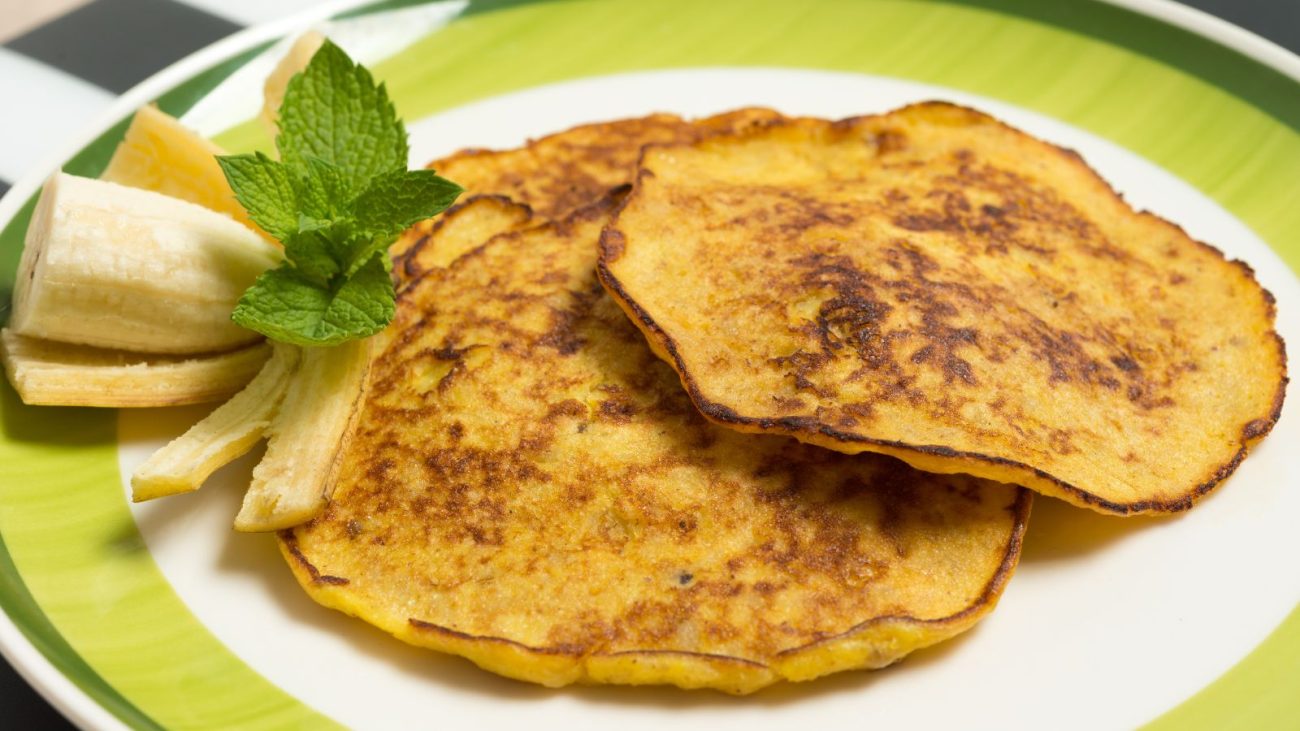 Panqueca sem farinha em um prato branco com decoração de hortelã e banana fatiada ao lado, ideal para café da manhã ou lanche.