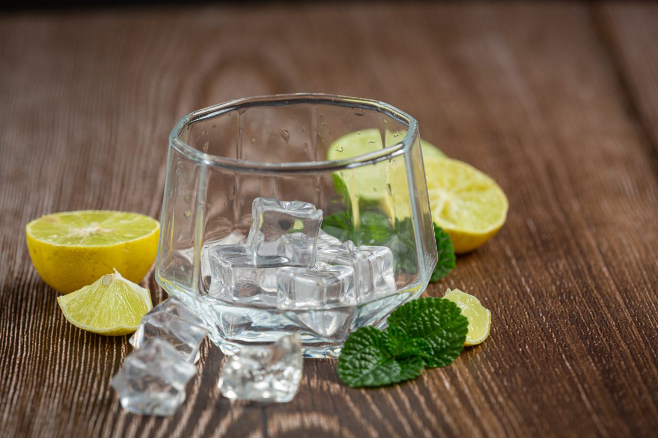 Copo de vidro transparente parcialmente preenchido com cubos de gelo. Ao redor do copo há pedaços de limão cortados e algumas folhas verdes de hortelã, tudo disposto sobre uma superfície de madeira rústica