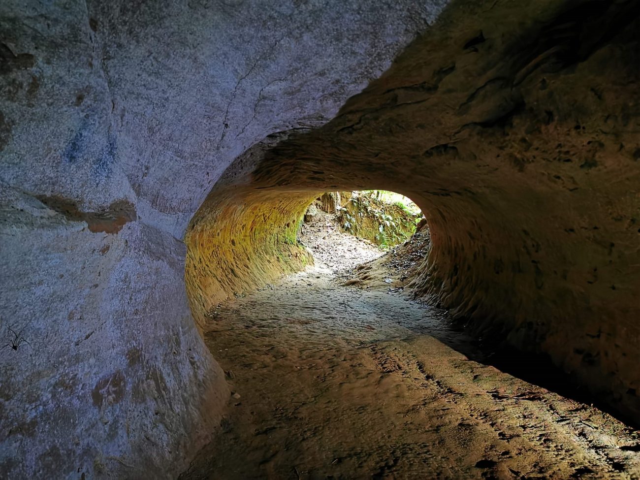 Foto de um grande vão dentro de uma rocha, por onde é possível passar pessoas. A luz do dia bate no vão e ilumina o caminho de terra.