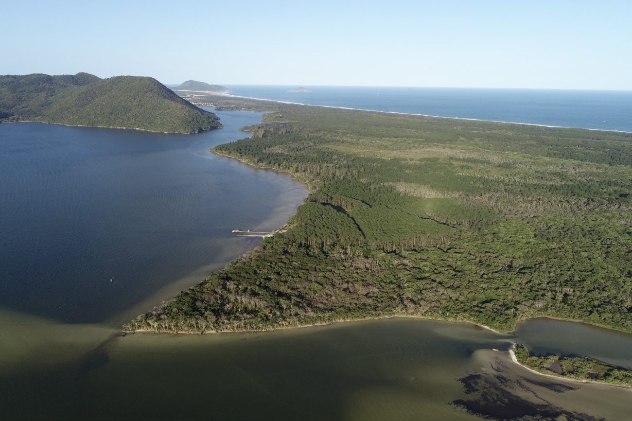 Vista aérea de uma ampla área verde cercada de um lado pelas águas da Lagoa da Conceição e de outro lado pelo mar. O céu está azul.