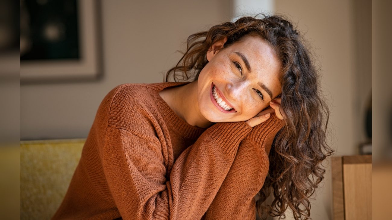 Mulher sorridente com cabelo cacheado e pele clara, usando suéter marrom, demonstrando charme e simpatia, que encanta pelo cisne signos que conquistam