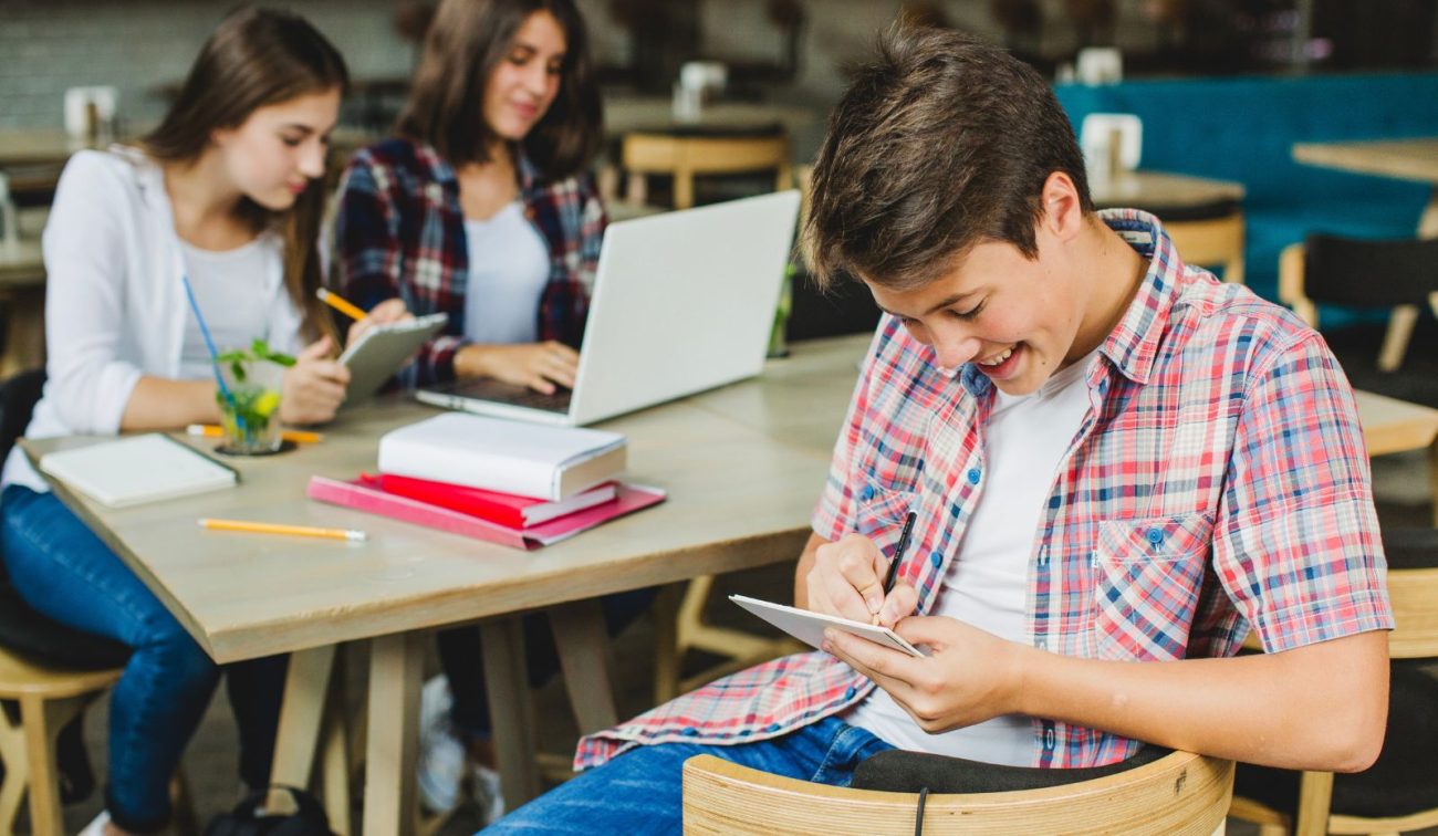 Três adolescentes estudam juntos em uma mesa; um deles utiliza o celular para anotações, representando o uso de celular em ambiente educacional.