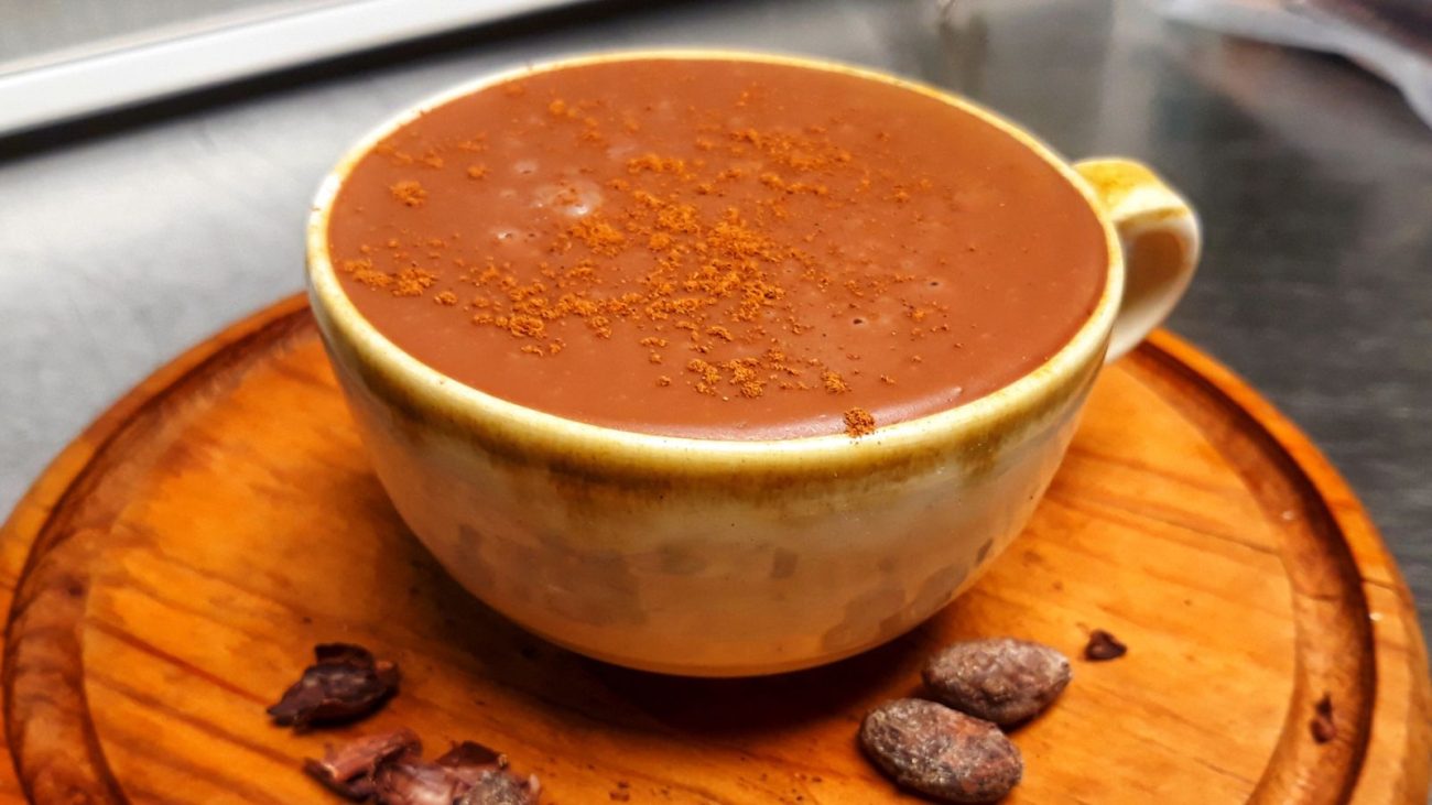 A imagem mostra uma xícara bege com detalhes marrons, cheia de chocolate quente cremoso e polvilhado com canela em pó por cima. A xícara está sobre um prato de madeira, e há alguns grãos e pedaços de cacau ao redor do prato.