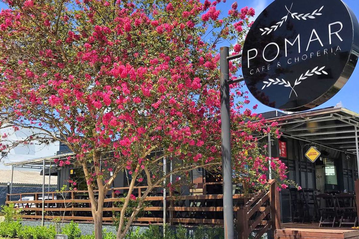 Em primeiro plano, uma árvore amplamente florida com flores rosa. Ao lado, uma placa circular preta com o nome Pomar Café e Choperia. Atrás, aparece parte do deck de madeira do estabelecimento, com algumas mesas também de madeira. 