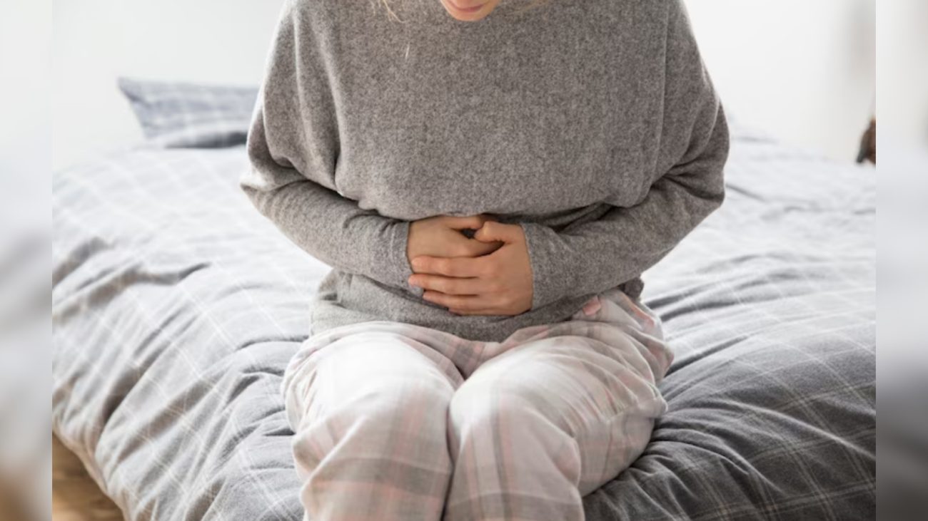 Pessoa sentada na beira de uma cama, usando pijama xadrez e blusa de moletom cinza. A pessoa está com as mãos pressionando a região abdominal inferior, com expressão de desconforto, indicando que pode estar sentindo dor abdominal ou cólica