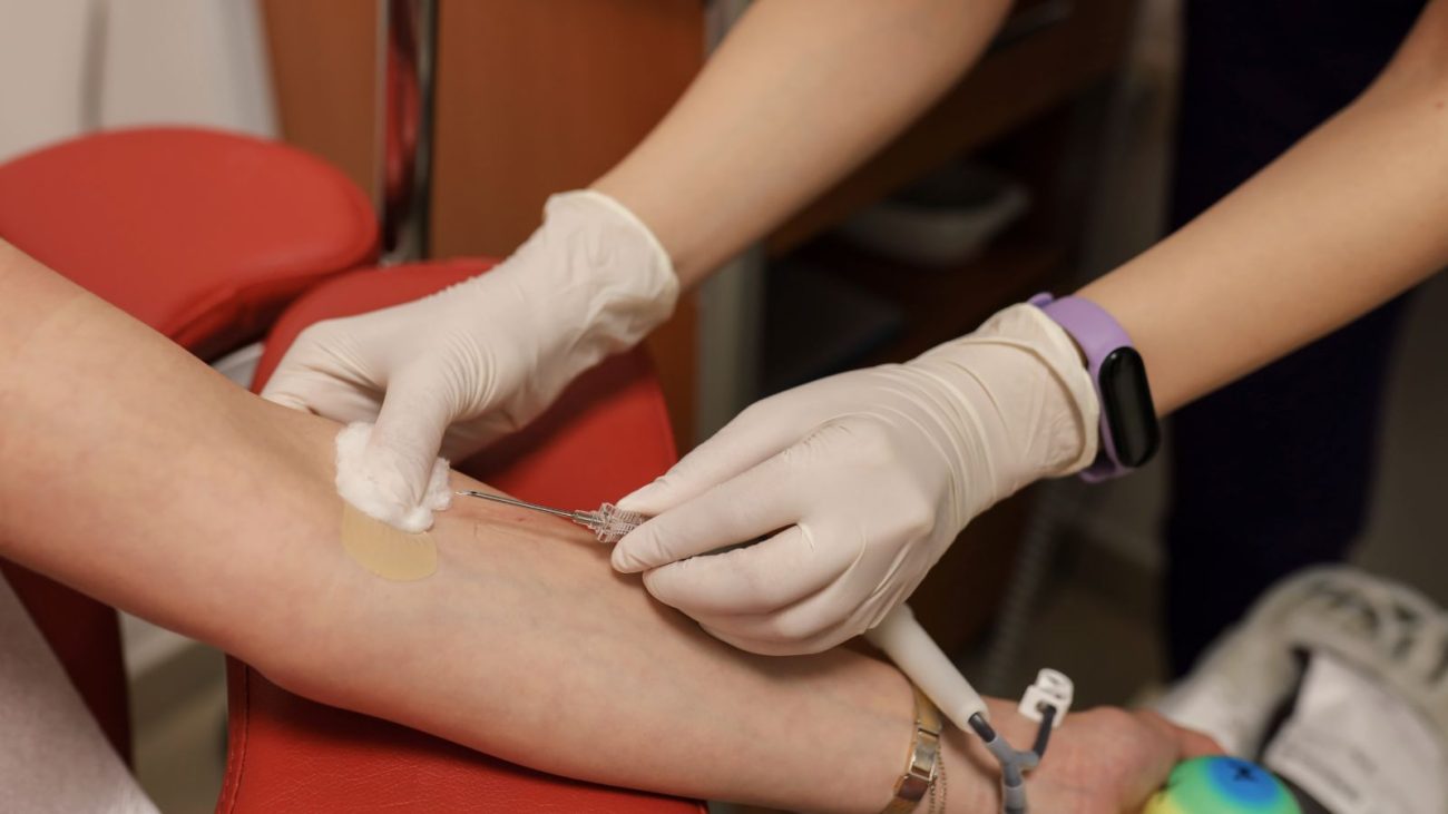 Imagem mostra um braço esticado, com uma agulha sendo colocada por duas mãos revestidas de luva cirúrgica branca para ilustrar como é doar sangue.