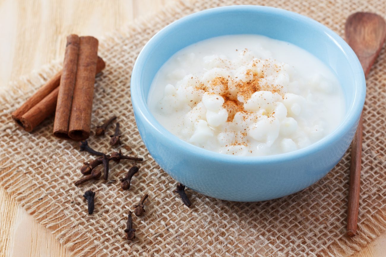Canjica cremosa para Festa Junina em um bowl azul, com pitadas de canela em cima, e pau de canela e cravo, uma colher de pau ao lado