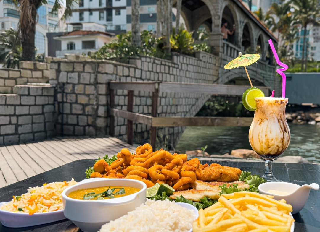 Foto de uma pequena mesa de madeira escura. Sobre ela, travessas com arroz, batata frita, pirão, peixe e camarão frito e uma taça de vidro com um drink. Ao fundo e desfocado, uma ponte de pedra e coberta passa sobre um rio.