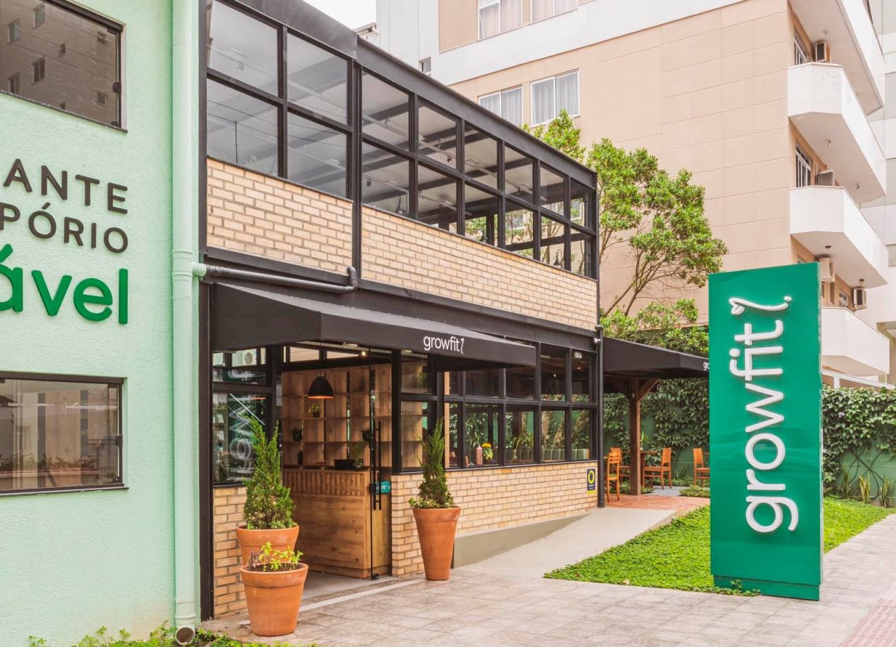 Foto lateral da fachada do Growfit, um dos restaurantes em Itapema. Na calçada, uma placa verde vertical com o nome do restaurante deitado. Ao lado, um prédio de tijolos à vista e muitos vidros quadrados. Na porta, três vasos grandes de barro com plantas.
