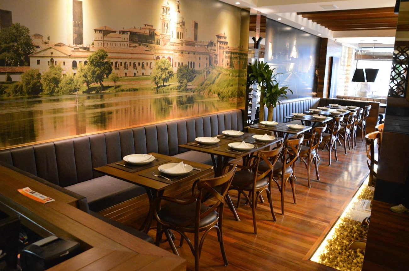 Salão interno de um dos restaurantes em Itapema tem várias pequenas mesas de madeira, com cadeiras de madeira de um lado e de outro lado um extenso sofá estofado em preto. O chão é laminado de madeira e nas paredes há grandes painéis de fotos.