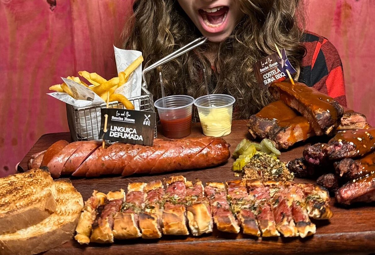 Foto de uma tábua de madeira com linguiça defumada cortada, pão de alho cortado em fatias, costela suína, uma cestinha com batata-fritas e ao dela pequenos potes de ketchup e maionese. Atrás da tábua, aparece em parte uma mulher de cabelos longos e a boca bem aberta, em uma expressão de alegria.
