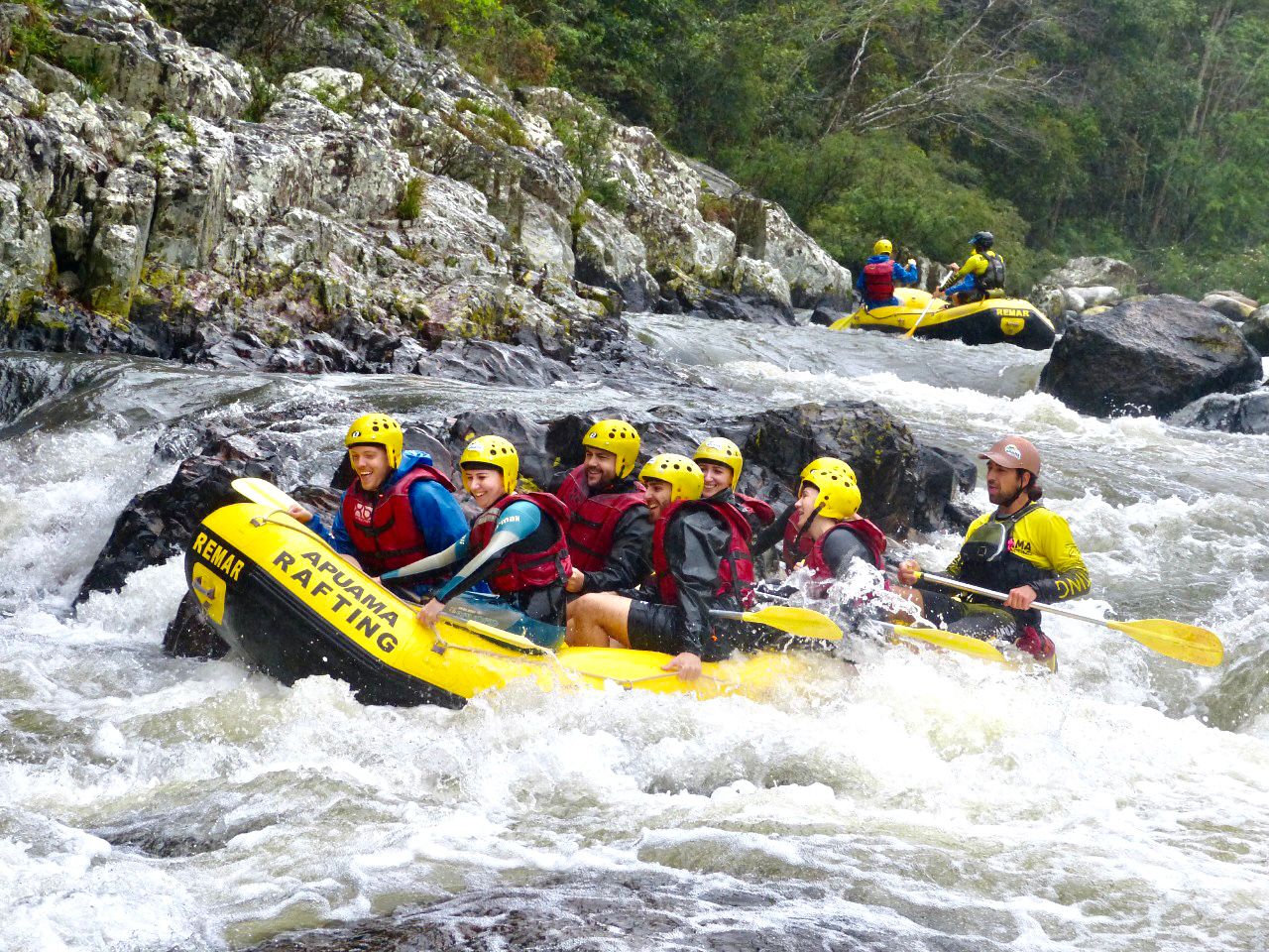 Foto de um grupo de oito pessoas em um bote inflável amarelo e preto. Eles estão dentro de um rio e passam por uma corredeira. A maioria das pessoas usa capacete e colete salva-vidas e estão sorrindo. Ao fundo, outro bote com duas pessoas, de costas para foto. No canto superior da foto, aparece parte de um paredão rochoso e vegetação.