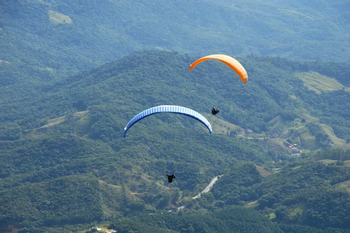 Foto de duas pessoas voando em dois parapentes em meio a grande morros verdes. Um dos parapente é azul e o outro é laranja.