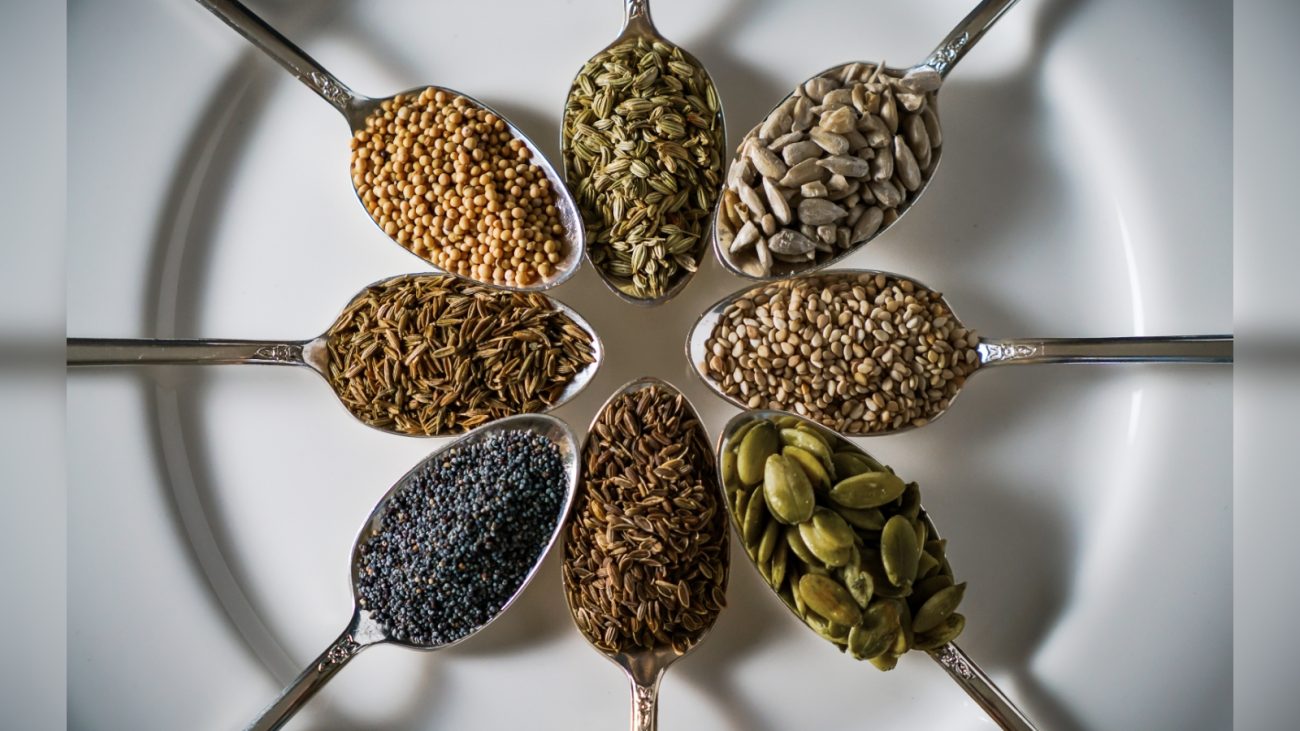 Diferentes sementes e especiarias organizadas em colheres dispostas em círculo, destacando variedade de ingredientes naturais e saudáveis.