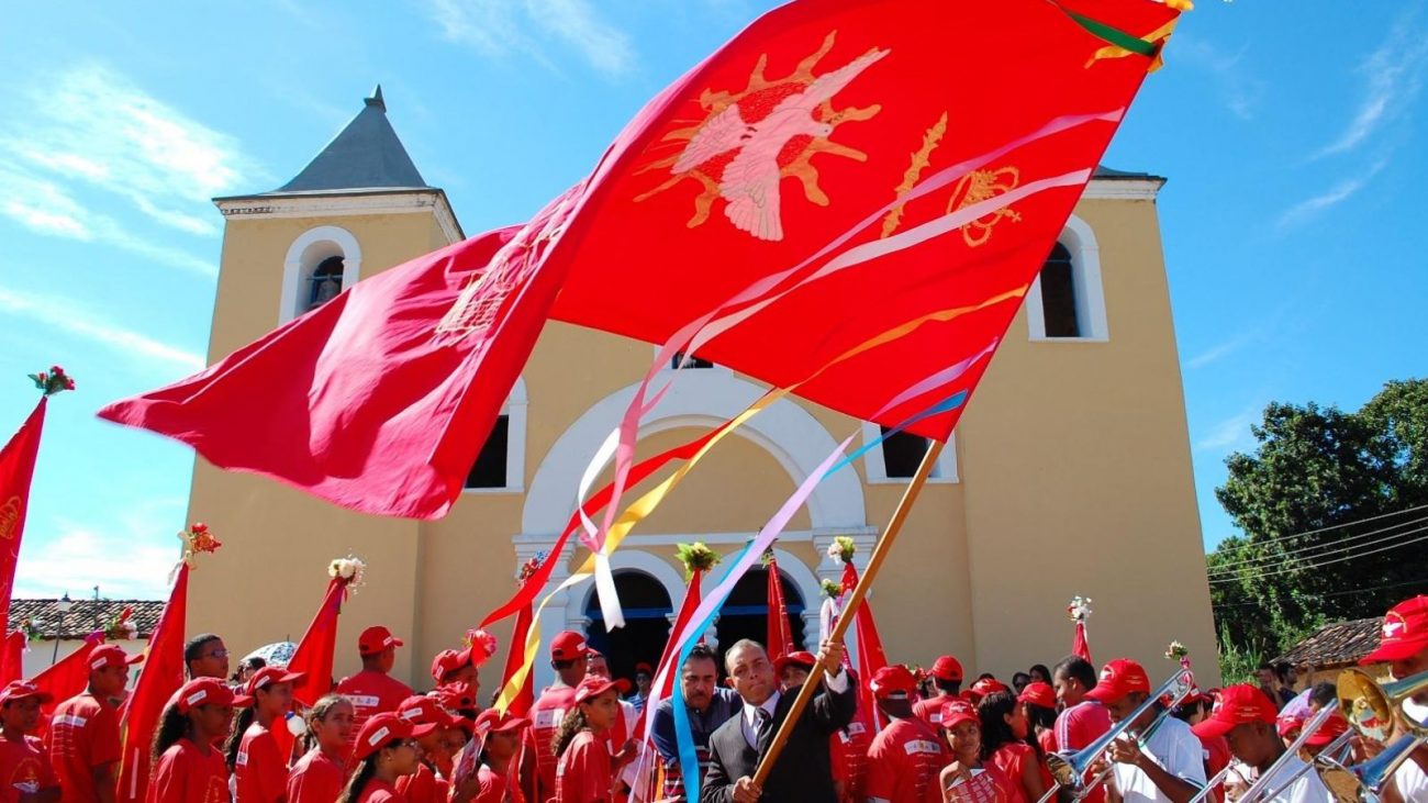 Imagem mostra as comemorações sendo realizadas em cidades de Tocantins para o Senhor do Bonfim para ilustrar o feriado que cai na sexta-feira em 2025. É possível ver uma multidão utilizando roupas vermelhar em frente a uma igreja católica amarela, com uma bandeira maior sendo carregada por um homem. Na bandeira tem um símbolo de uma pomba branca com asas abertas. 