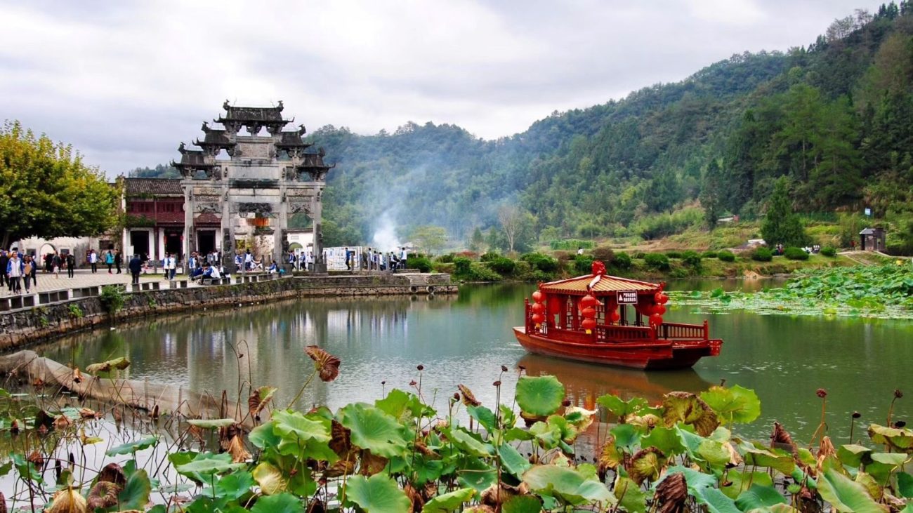 Paisagem tradicional na China com um lago, um barco vermelho, arquitetura antiga e uma ponte, rodeado por montanhas e vegetação exuberante.