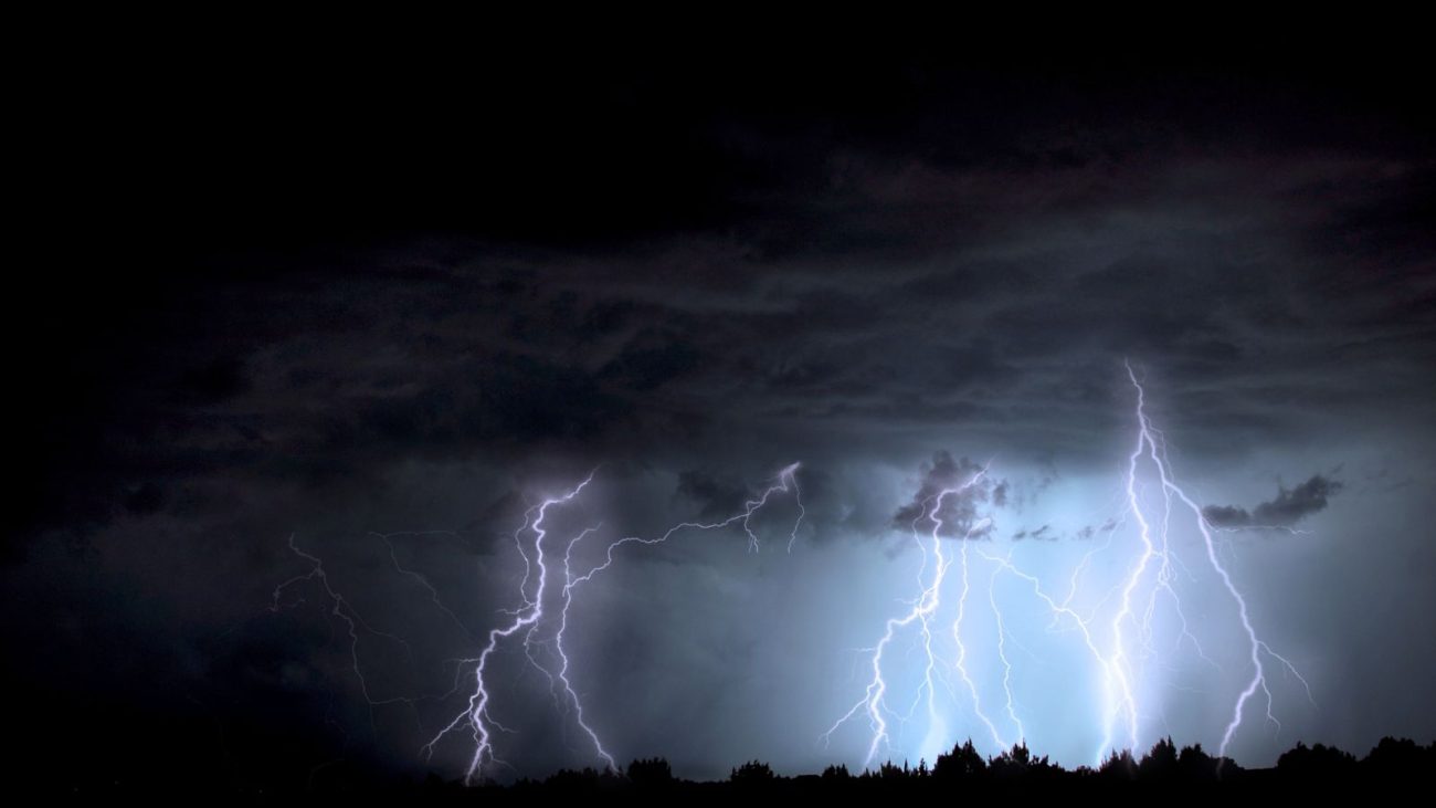 Céu noturno carregado por nuvens escuras e intensos raios durante uma forte tempestade, evidenciando a chegada de uma frente fria. A imagem mostra múltiplos relâmpagos iluminando a paisagem, com silhuetas de árvores ao fundo.