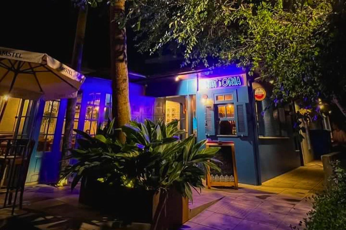 Casa com paredes azuis e um letreiro escrito Tratoria. Na frente e na lateral dela, aparecem partes de árvores. 