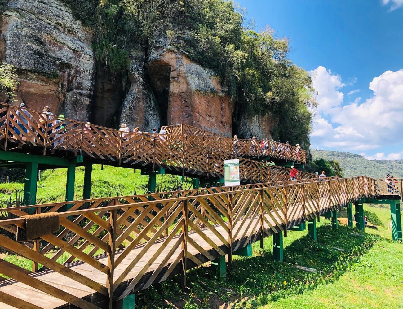 Em primeiro plano, uma extensa passarela de madeira, sobre a qual caminham dezenas de pessoas. Ao fundo, um grande paredão rochoso com vegetações. O céu está azul, com algumas nuvens.