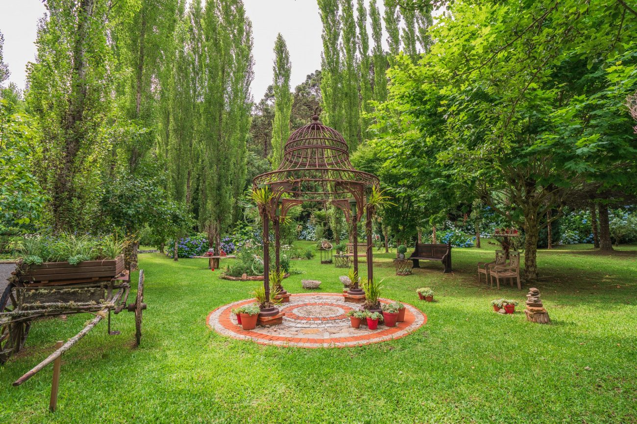 Foto de uma ampla área gramada com árvores de diferentes tipos e tamanhos e alguns bancos de madeira. No centro da foto, há uma estrutura decorativa, alta e de aço. Ao redor dela, vasos com flores.