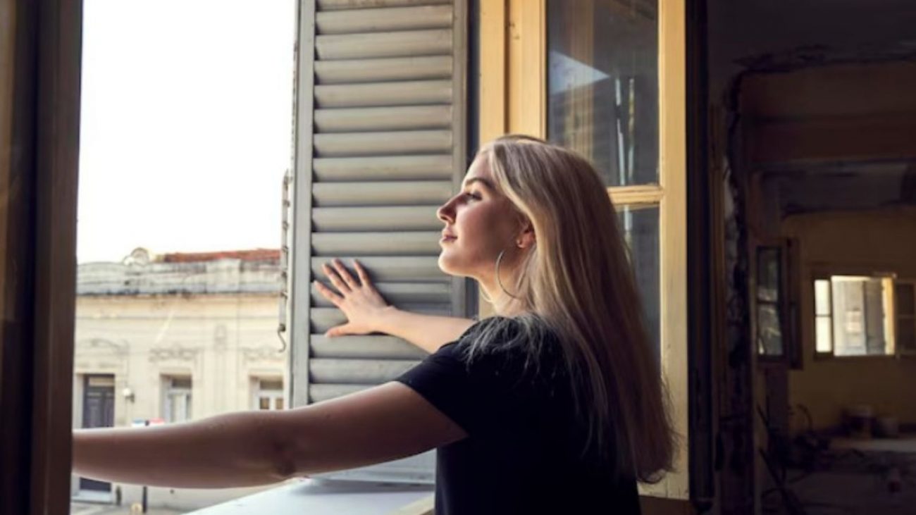 na foto aparece uma mulher que abre a janela para ventilar a casa todos os dias
