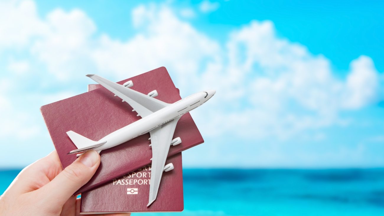 "Mão segurando dois passaportes de capa vermelha com a palavra 'PASSPORT' escrita em branco. Sobre os passaportes, há uma miniatura de avião branco. Ao fundo, céu azul com nuvens e o mar em tom turquesa, em um cenário que remete a viagens e férias