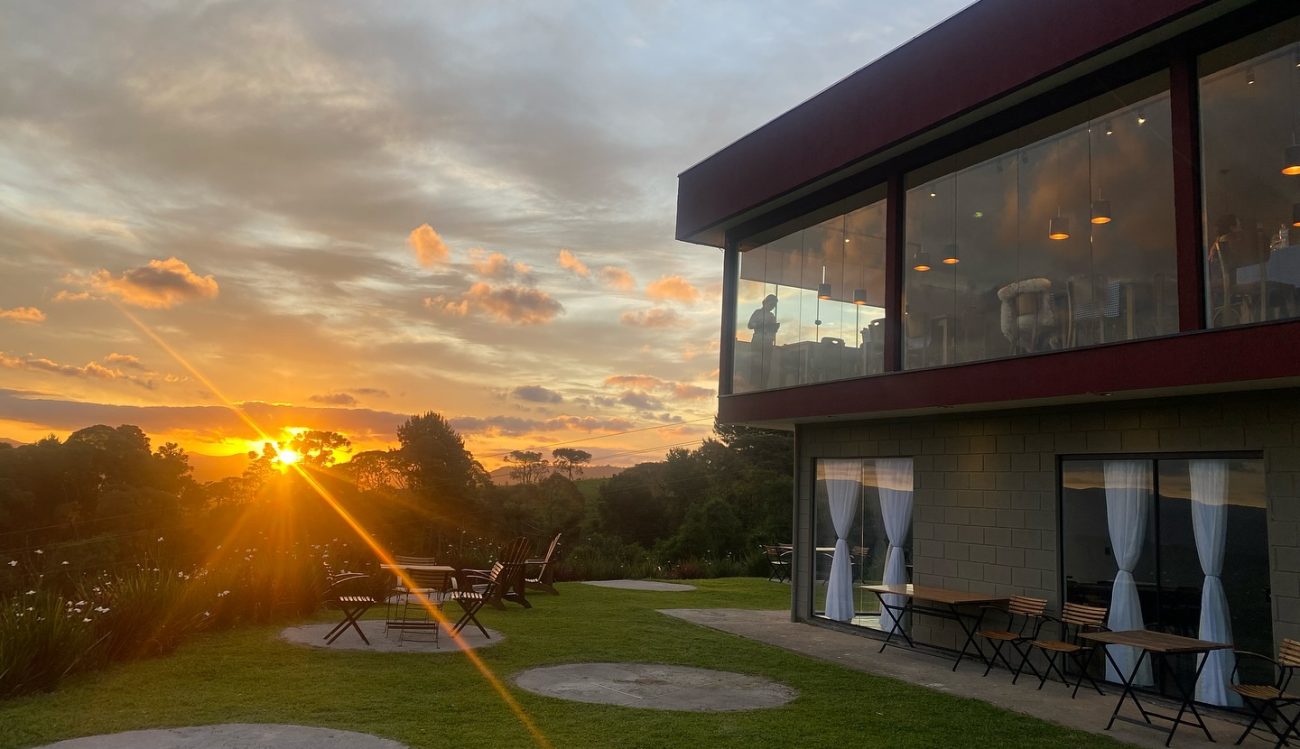 No lado direito, um prédio de dois andares com grandes janelas de vidro da Vinícola Serra do Sol. À esquerda, um gramado com algumas mesas e cadeiras de madeira. Ao fundo, o pôr do sol e o céu azul alaranjado.
