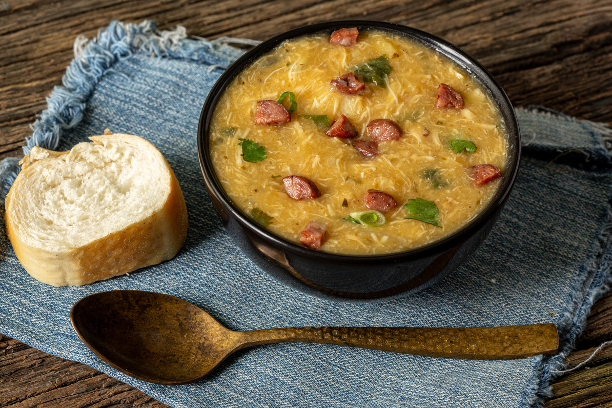 Caldo de Kenga, que é um caldo de mandioca com frango desfiado e calabresa, em panela rústica sobre guradanapo de pano e fatia de pão e colher de ferro ao lado 