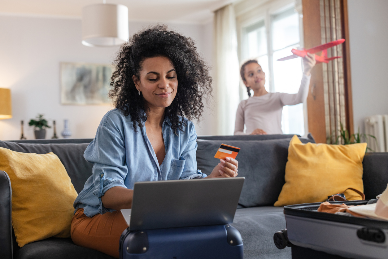 Mulher sentada no sofá com cartão de banco em uma mão e a outra mexendo no notebook. Ao fundo, criança brinca com avião