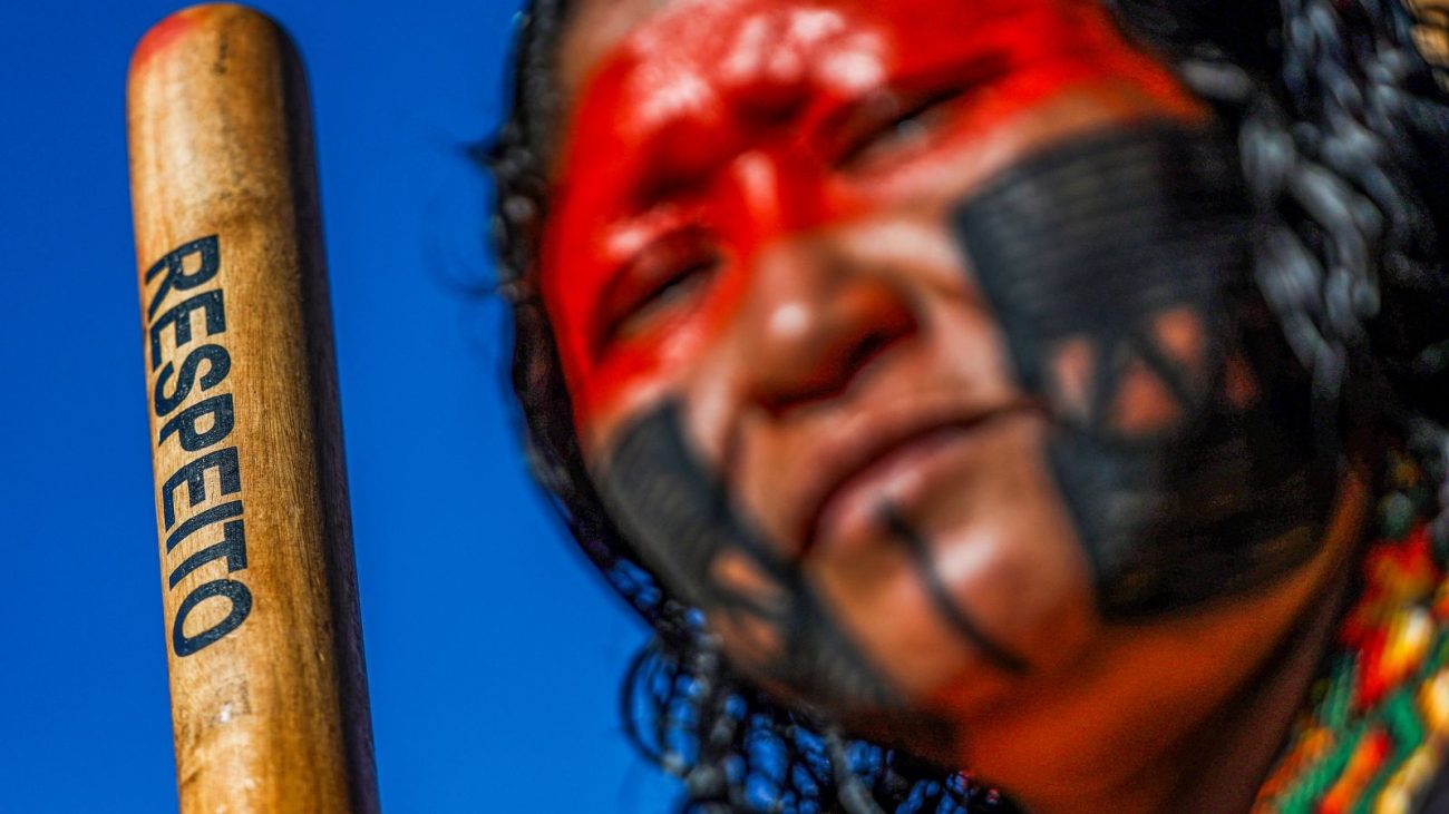 Mulher indígena com rosto pintado em vermelho e preto segura um bastão com a palavra “respeito” escrita. A imagem simboliza a luta dos povos originários contra ações ilegais como a introdução de dispositivos de áudio em áreas protegidas da Amazônia.