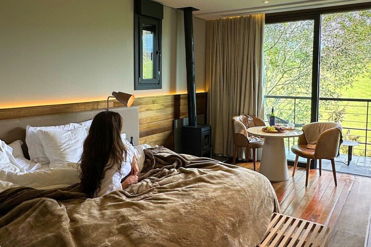 Foto de uma mulher jovem de cabelos castanhos longos deitada em uma grande cama de casal com cobertor marrom, dentro do quarto da pousada. Ela olha para fora, onde aparece parte de uma árvore e um campo verde. Ao lado da cama, uma lareira preta e uma pequena mesa com duas cadeiras. 