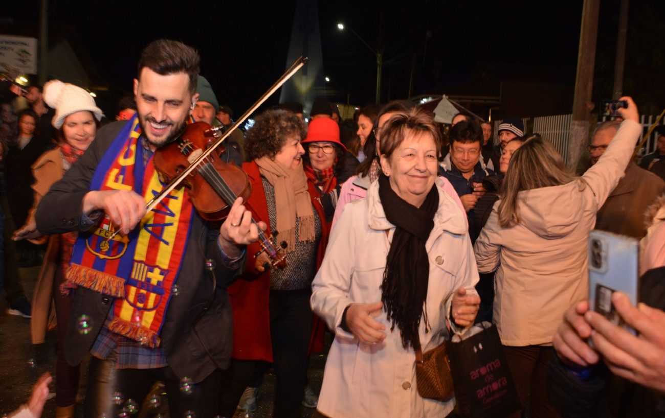 Foto de um homem jovem sorrindo e tocando violino na rua. Ao lado dele, uma senhora com casaco e cachecol sorri. Atrás deles, dezenas de pessoas com roupas de frio sorriem e tiram fotos. 