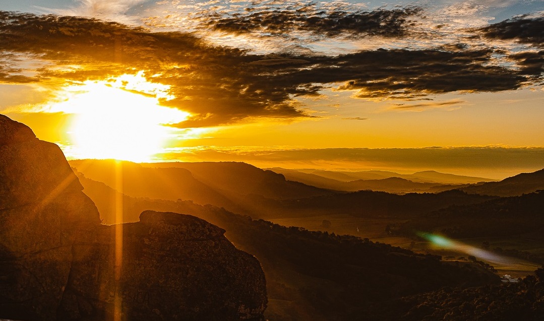 Foto horizontal. Em primeiro plano, aparecem morros escuros, em contraluz. Por detrás deles, o sol se põe. Alguns raios luminosos batem nos morros. O céu tem algumas nuvens e está alaranjado. 