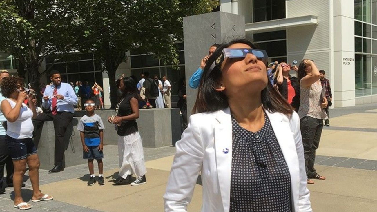 Mulher usando óculos de realidade virtual em um ambiente ao ar livre, com pessoas ao fundo na cidade, participando de uma experiência imersiva.