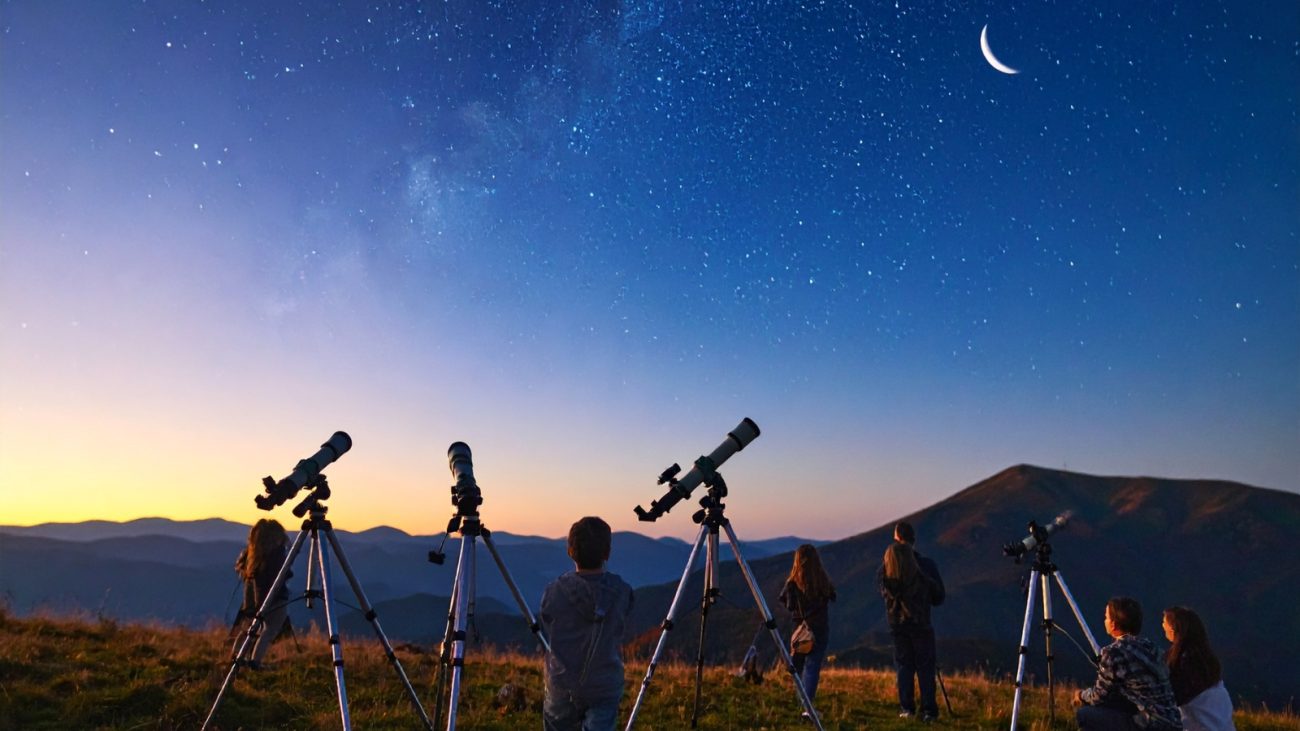 Grupo de pessoas observando o céu noturno com telescópios em uma paisagem de montanhas ao entardecer, com a Lua visível no céu estrelado